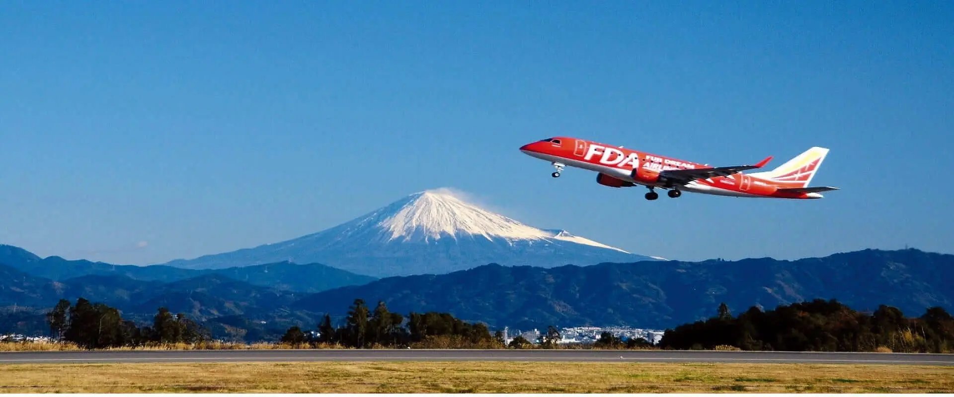 雪をかぶった富士山を背景に青空へ飛び立つ赤いFDAの飛行機